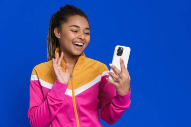 Siyah genç bir kadın mavi arka planda cep telefonuyla selfie çekerken el kol hareketi yapıyor.