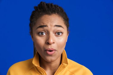 Siyahi genç kadın şaşkınlığını dile getiriyor ve mavi arka planda izole edilmiş kameraya bakıyor.