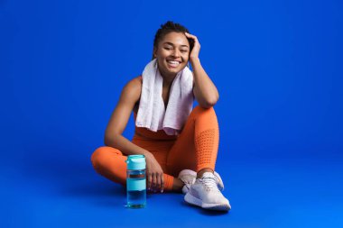 Mavi arka planda izole bir şekilde yerde otururken gülümseyen genç zenci sporcu kadın.