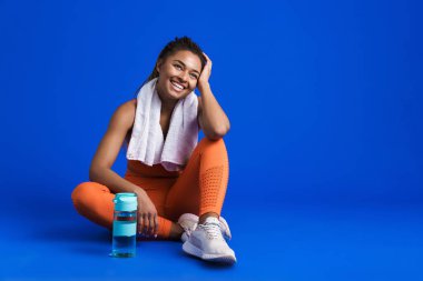 Mavi arka planda izole bir şekilde yerde otururken gülümseyen genç zenci sporcu kadın.