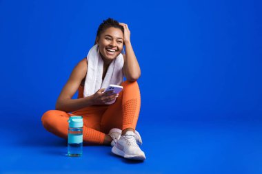 Mavi arka planda izole bir şekilde yerde otururken cep telefonu kullanan siyahi genç sporcu.