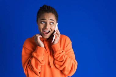 Mavi arka planda izole bir şekilde cep telefonuyla konuşurken şaşırdığını ifade eden siyahi genç bir kadın.