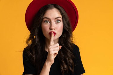 White shocked woman in red hat showing silence gesture isolated over yellow background
