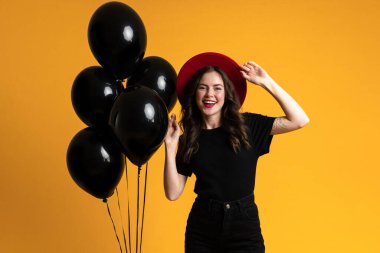 Kırmızı şapkalı beyaz kadın, sarı arka planda siyah balonlarla poz verirken gülümsüyor.