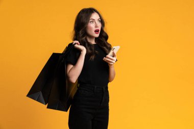 Alış veriş torbaları ve cep telefonuyla poz veren beyaz kadın sarı arka planda izole edilmiş.