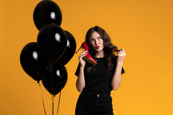 White woman holding high heel while posing with black balloons isolated over yellow background