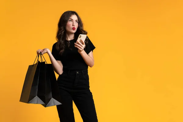 White Puzzlement Woman Posing Shopping Bags Cellphone Isolated Yellow Background — Stockfoto