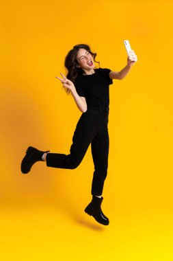 Beyaz heyecanlı kadın sarı arka planda izole edilmiş cep telefonuyla selfie çekerken el kol hareketi yapıyor.