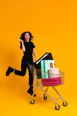 Beyaz heyecanlı kadın, sarı arka planda izole edilmiş alışveriş arabasıyla poz verirken gülüyor.