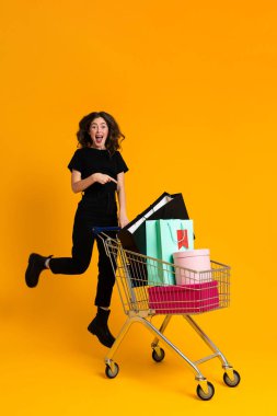 White excited woman pointing finger at shopping cart isolated over yellow background
