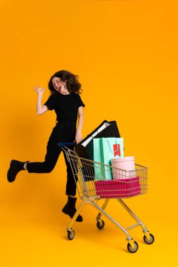 White excited woman laughing while posing with shopping cart isolated over yellow background