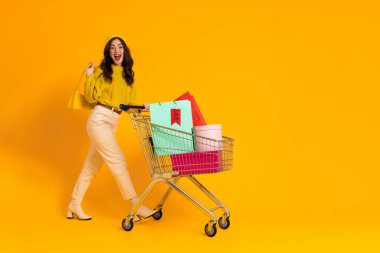 White excited woman laughing while posing with shopping cart isolated over yellow background