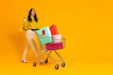 White excited woman laughing while posing with shopping cart isolated over yellow background