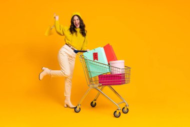 White excited woman laughing while posing with shopping cart isolated over yellow background