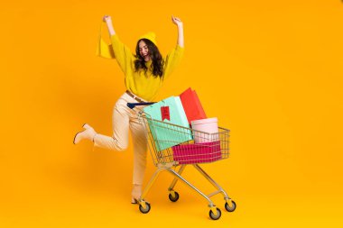 White woman making winner gesture while posing with shopping cart isolated over yellow background