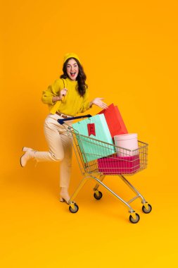 White excited woman laughing while posing with shopping cart isolated over yellow background