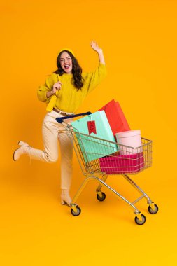 White woman making fun while posing with shopping cart isolated over yellow background