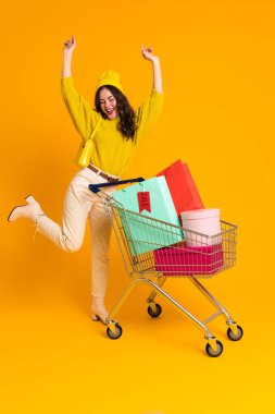 White excited woman making fun while posing with shopping cart isolated over yellow background