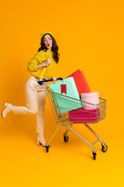 White excited woman grimacing while posing with shopping cart isolated over yellow background