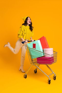 White excited woman laughing while posing with shopping cart isolated over yellow background