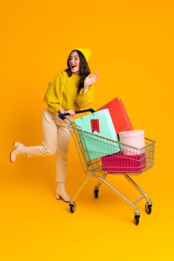 White excited woman gesturing while posing with shopping cart isolated over yellow background