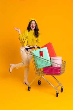 White woman holding copyspace while posing with shopping cart isolated over yellow background
