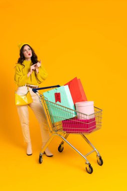 Sarı arka planda izole edilmiş alışveriş arabasıyla poz veren beyaz şaşkın kadın.