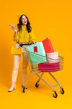 White excited woman posing with cellphone and shopping cart isolated over yellow background