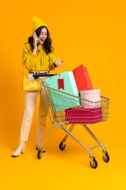 Excited woman talking on cellphone while posing with shopping cart isolated over yellow background