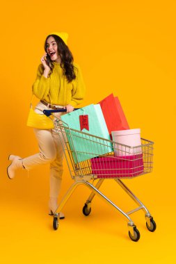 Excited woman talking on cellphone while posing with shopping cart isolated over yellow background