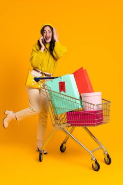 Excited woman talking on cellphone while posing with shopping cart isolated over yellow background