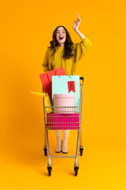 White excited woman gesturing while posing with shopping cart isolated over yellow background
