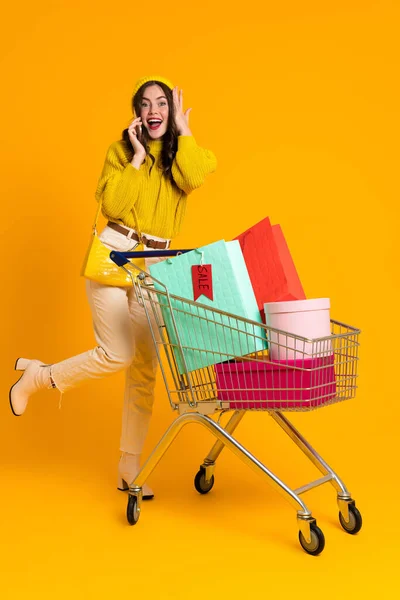 Excited Woman Talking Cellphone While Posing Shopping Cart Isolated Yellow — Φωτογραφία Αρχείου