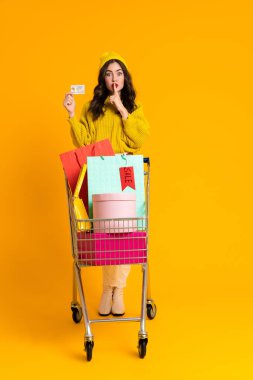 White woman posing with credit card and shopping cart isolated over yellow background