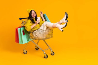White excited woman laughing and using cellphone in shopping cart isolated over yellow background