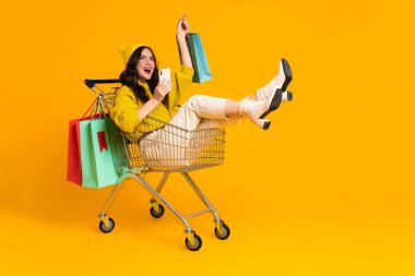 White excited woman laughing and using cellphone in shopping cart isolated over yellow background