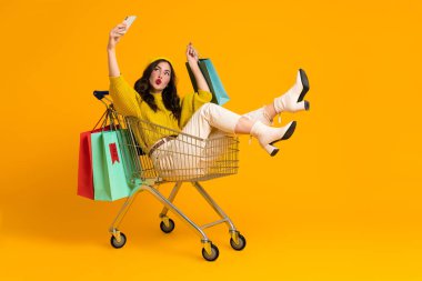 White young woman taking selfie on cellphone in shopping cart isolated over yellow background