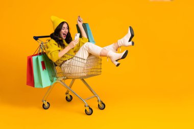 White excited woman laughing and using cellphone in shopping cart isolated over yellow background