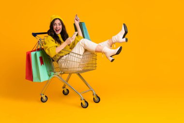 White excited woman laughing and using cellphone in shopping cart isolated over yellow background