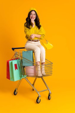 White woman smiling while sitting in shopping cart isolated over yellow background