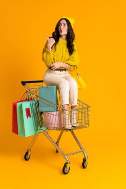 White woman pointing finger aside while sitting in shopping cart isolated over yellow background