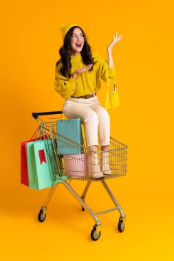 White woman holding copyspace while sitting in shopping cart isolated over yellow background