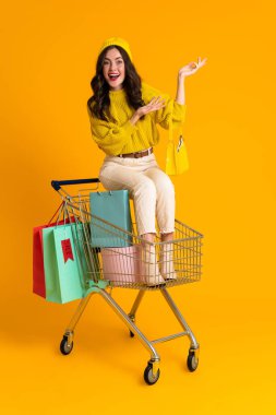 White woman holding copyspace while sitting in shopping cart isolated over yellow background