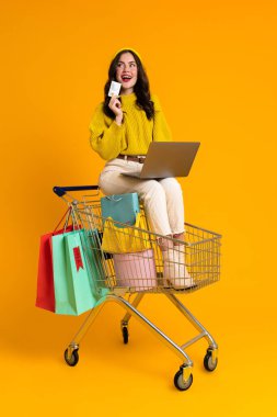 White woman using laptop while sitting in shopping cart isolated over yellow background