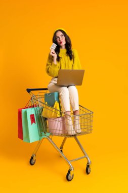 White woman using laptop while sitting in shopping cart isolated over yellow background