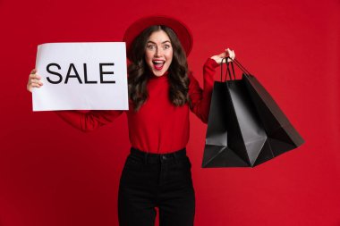 White excited woman posing with shopping bags and sale placard isolated over red background
