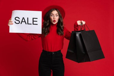 White surprised woman posing with shopping bags and sale placard isolated over red background