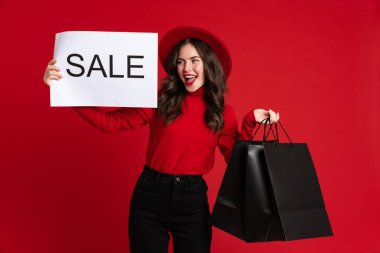 White woman smiling while posing with shopping bags and sale placard isolated over red background