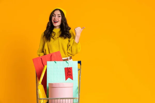Excited Woman Pointing Finger Aside While Posing Shopping Cart Isolated — стоковое фото