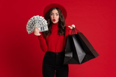 White woman grimacing while posing with shopping bags and dollars isolated over red background